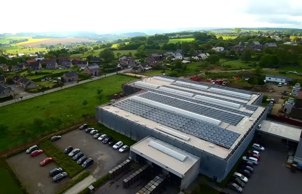 Vue aérienne d'un bâtiment industriel couvert de panneaux solaires, entouré de prairies et de maisons