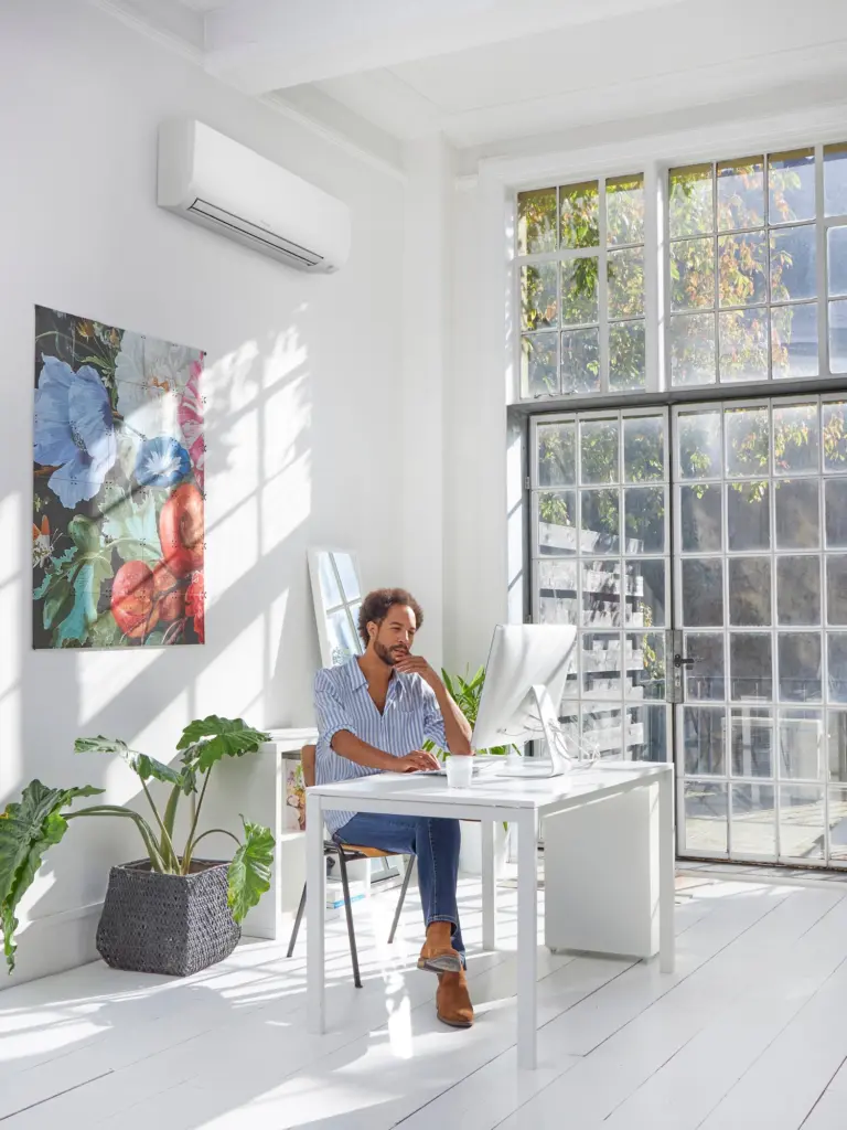Climatiseur mural Daikin Comfora blanc en haut d’un mur, dans un bureau lumineux aux grandes fenêtres